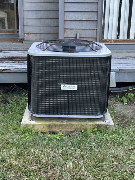 A large air conditioner is sitting in the grass in front of a house.