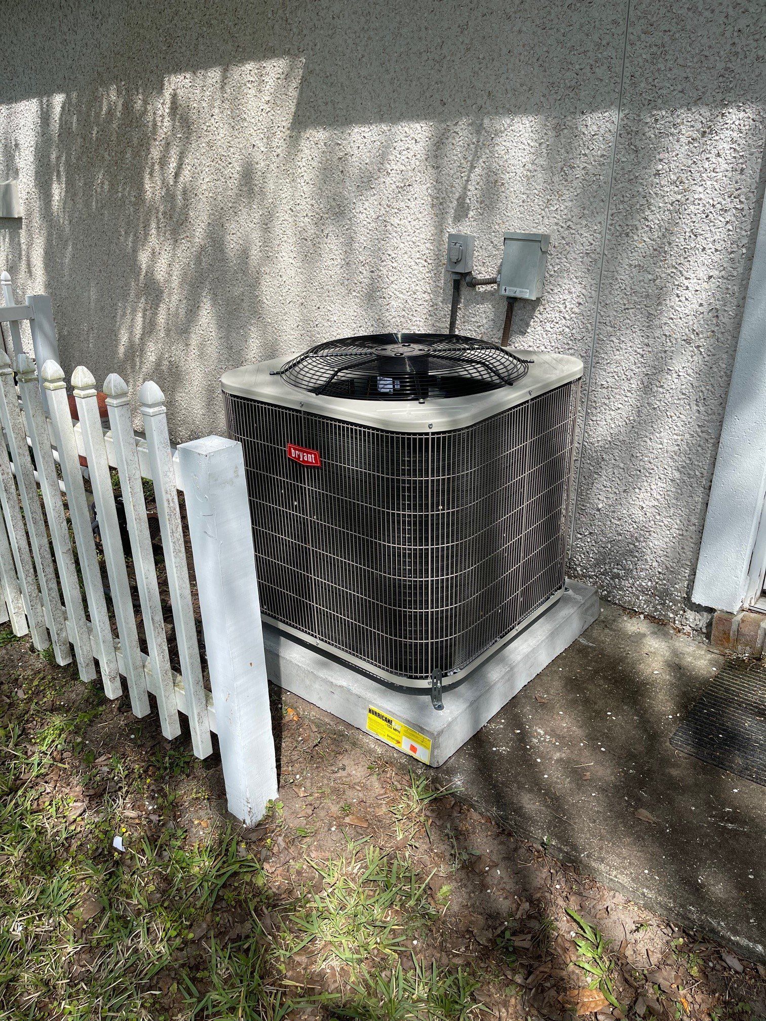 A black air conditioner is sitting on the side of a building next to a white picket fence.