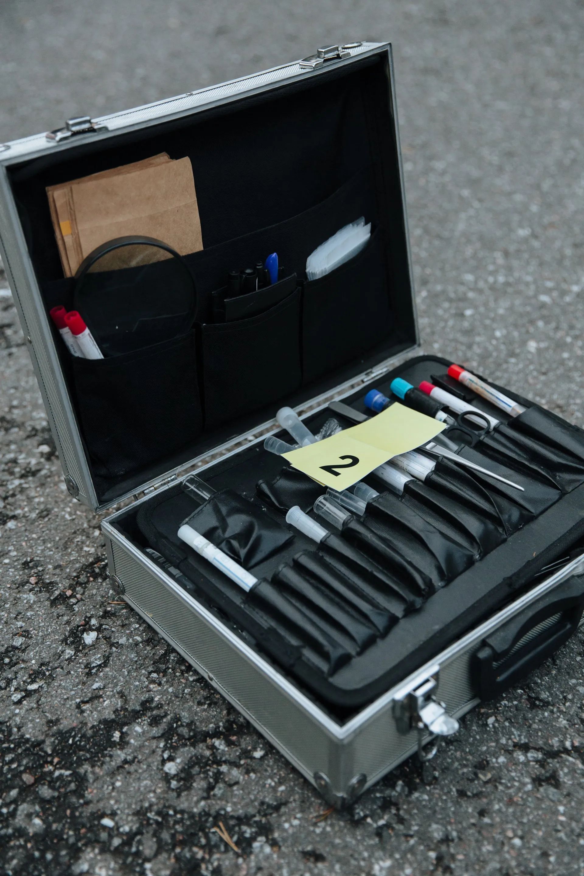Open silver case on asphalt with forensic tools, including brushes and vials.