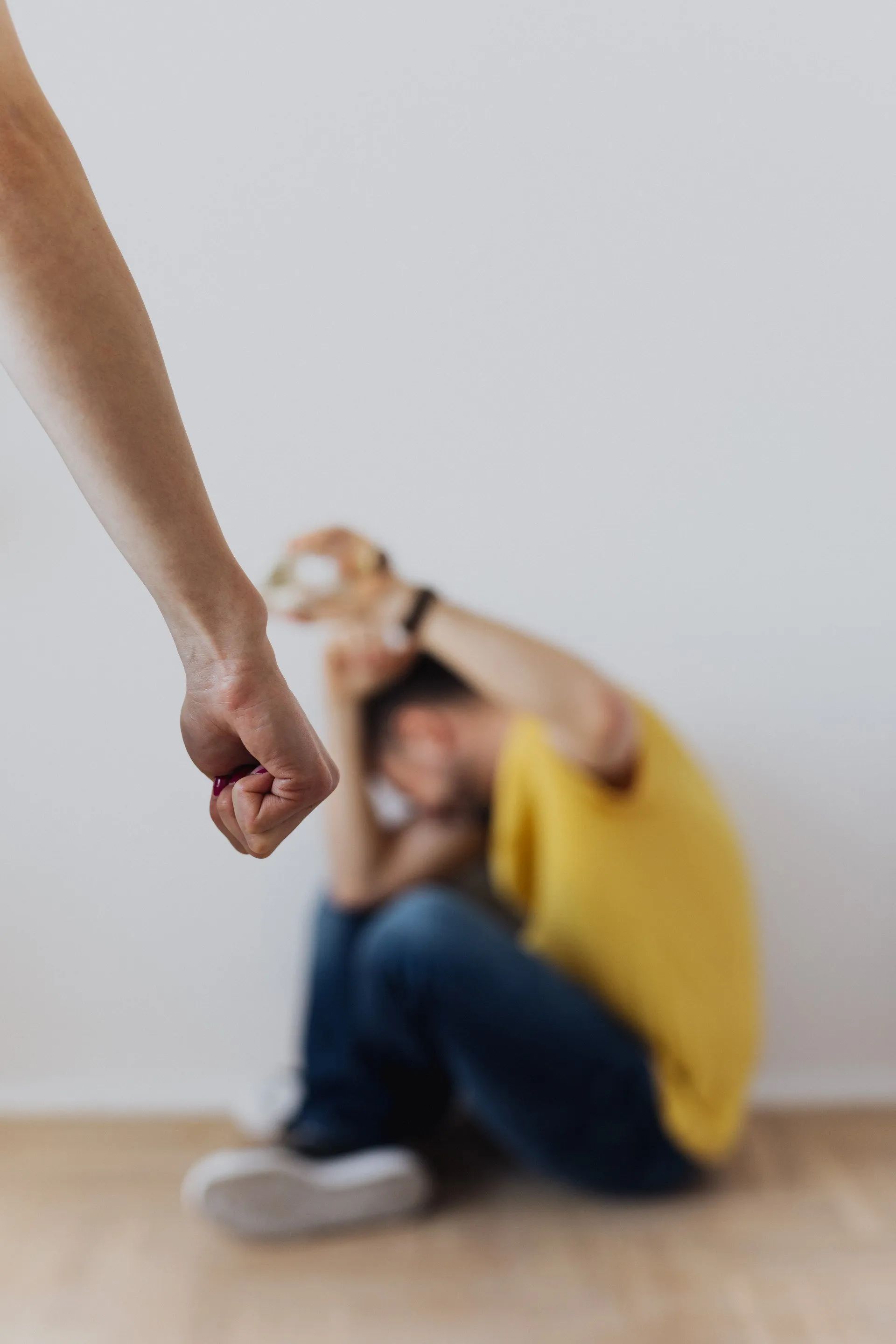 Person's fist raised toward another person cowering on the floor with hands over head.