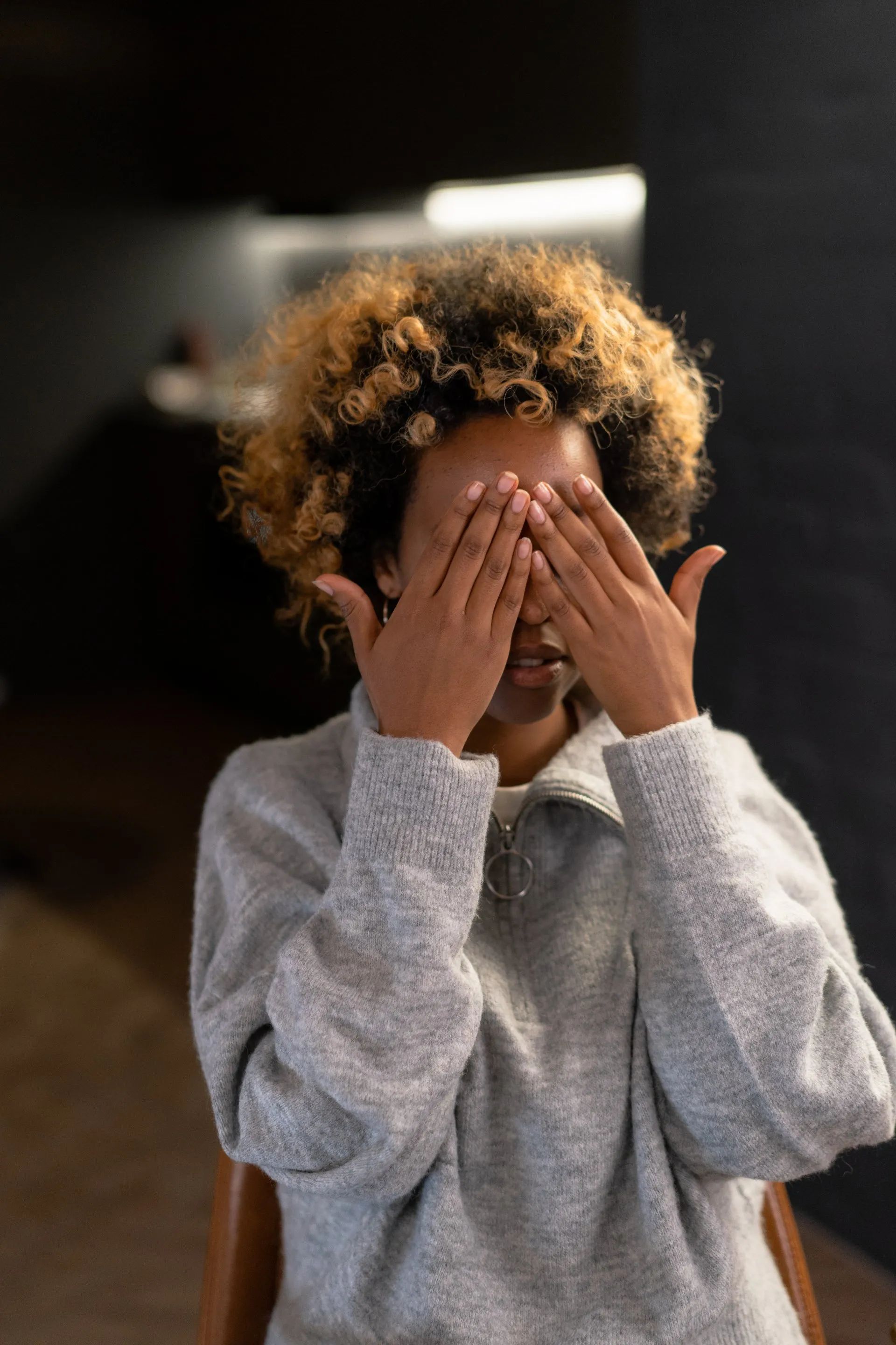 Person with curly hair covering their eyes with both hands. Wearing a grey sweater. Dark background.