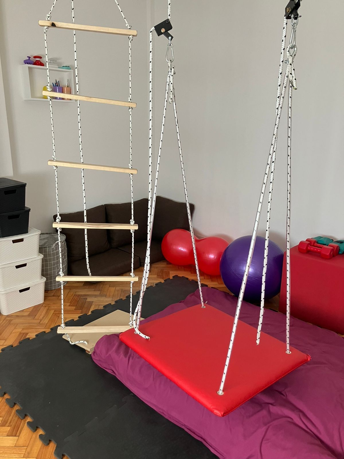 Un columpio terapéutico de interior con una plataforma roja y una escalera de cuerda que cuelga sobre una colchoneta.