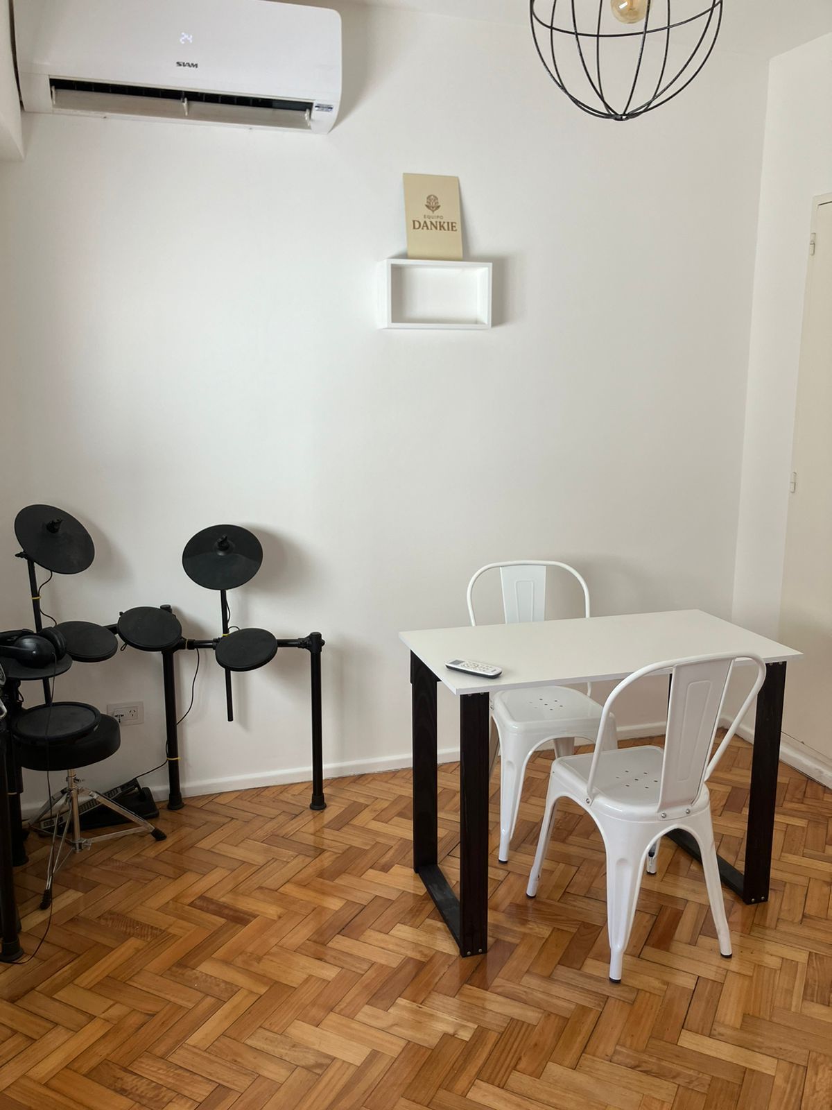 Sobre un suelo de parqué de madera, una mesa blanca con dos sillas de metal blancas y una batería electrónica en una esquina.
