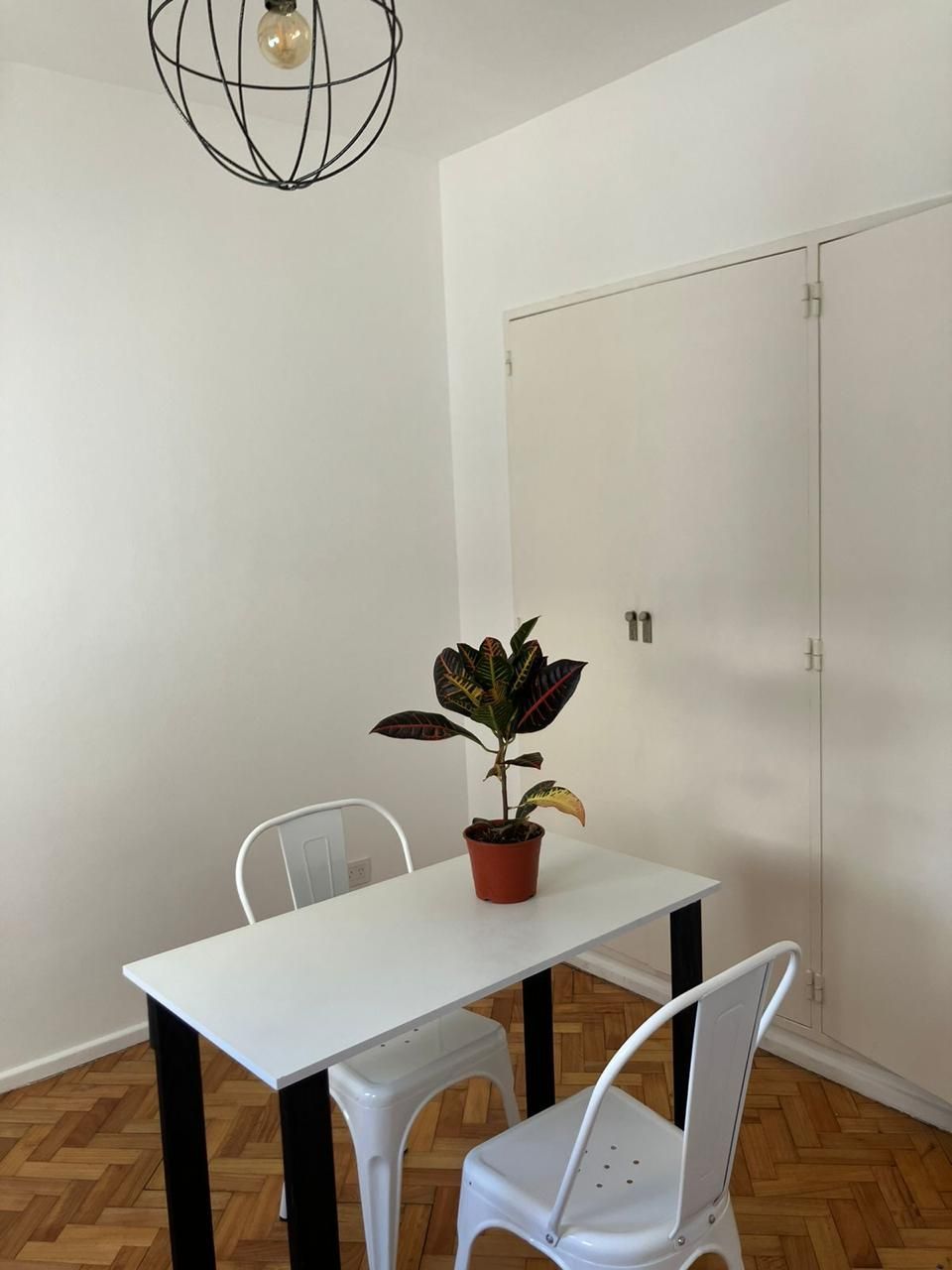 Sobre un suelo de parqué de madera, hay una mesa rectangular blanca con dos sillas de metal blancas y una planta en maceta.