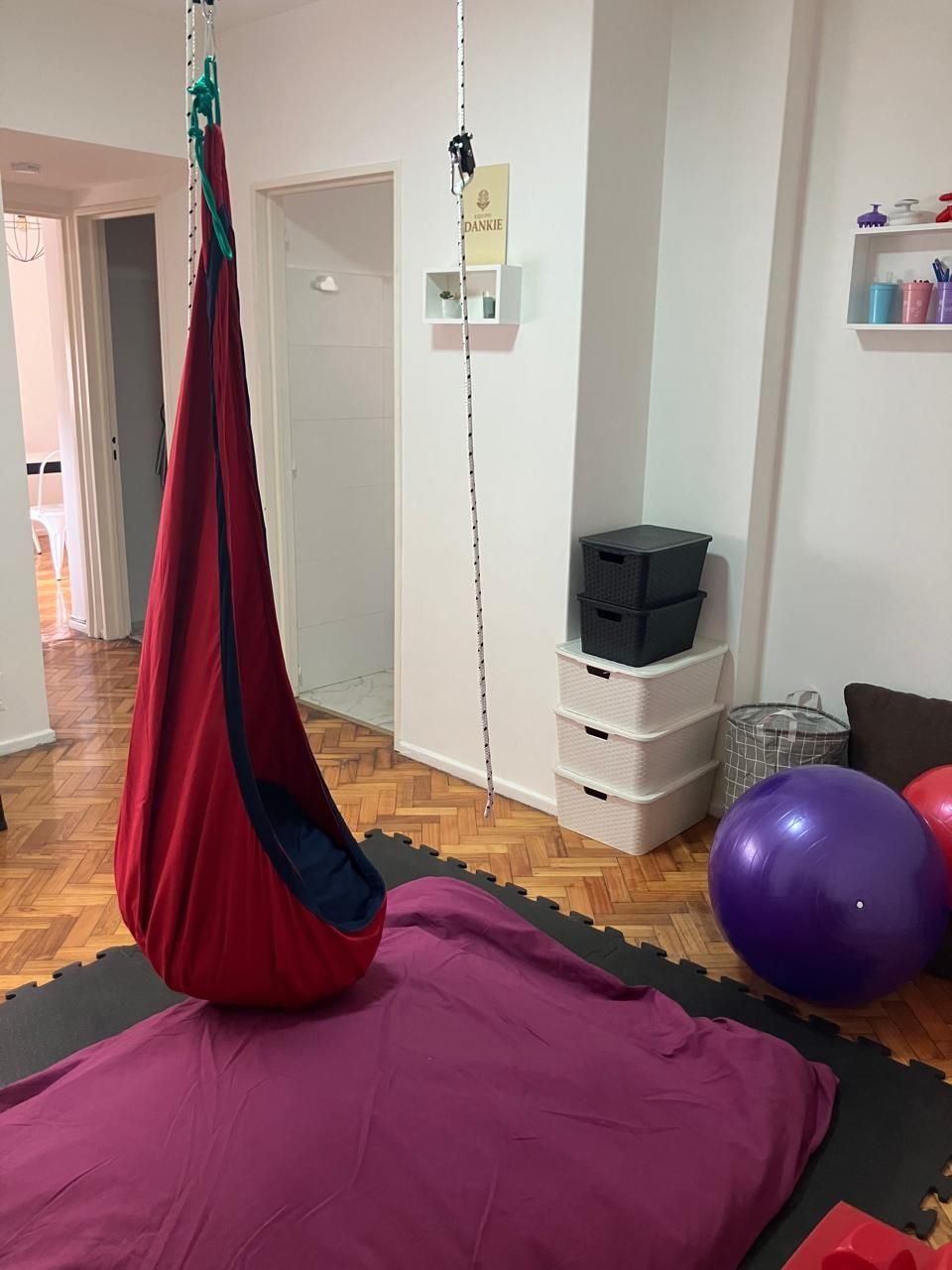 Un columpio sensorial rojo cuelga sobre una alfombra morada en una habitación con una pelota de yoga.