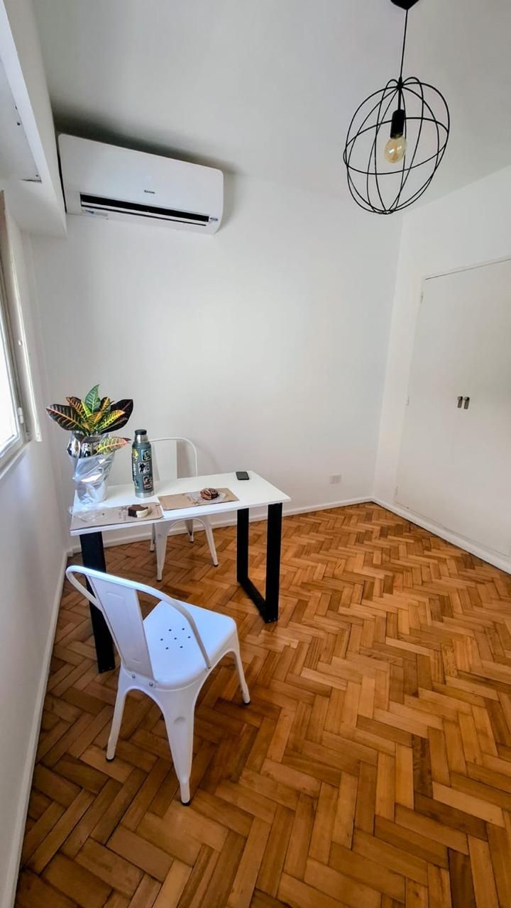 Un escritorio blanco con una silla de metal y una planta sobre un suelo de madera, bajo una lámpara colgante geométrica.