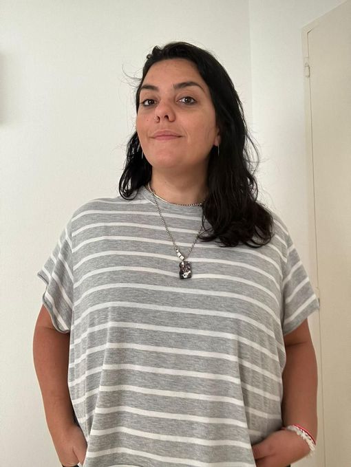 Una persona que lleva una camiseta de rayas grises y blancas y un collar con colgante, de pie frente a una pared blanca lisa.