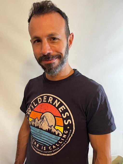 Una persona con barba, que lleva una camiseta azul marino con el dibujo de una montaña al atardecer.