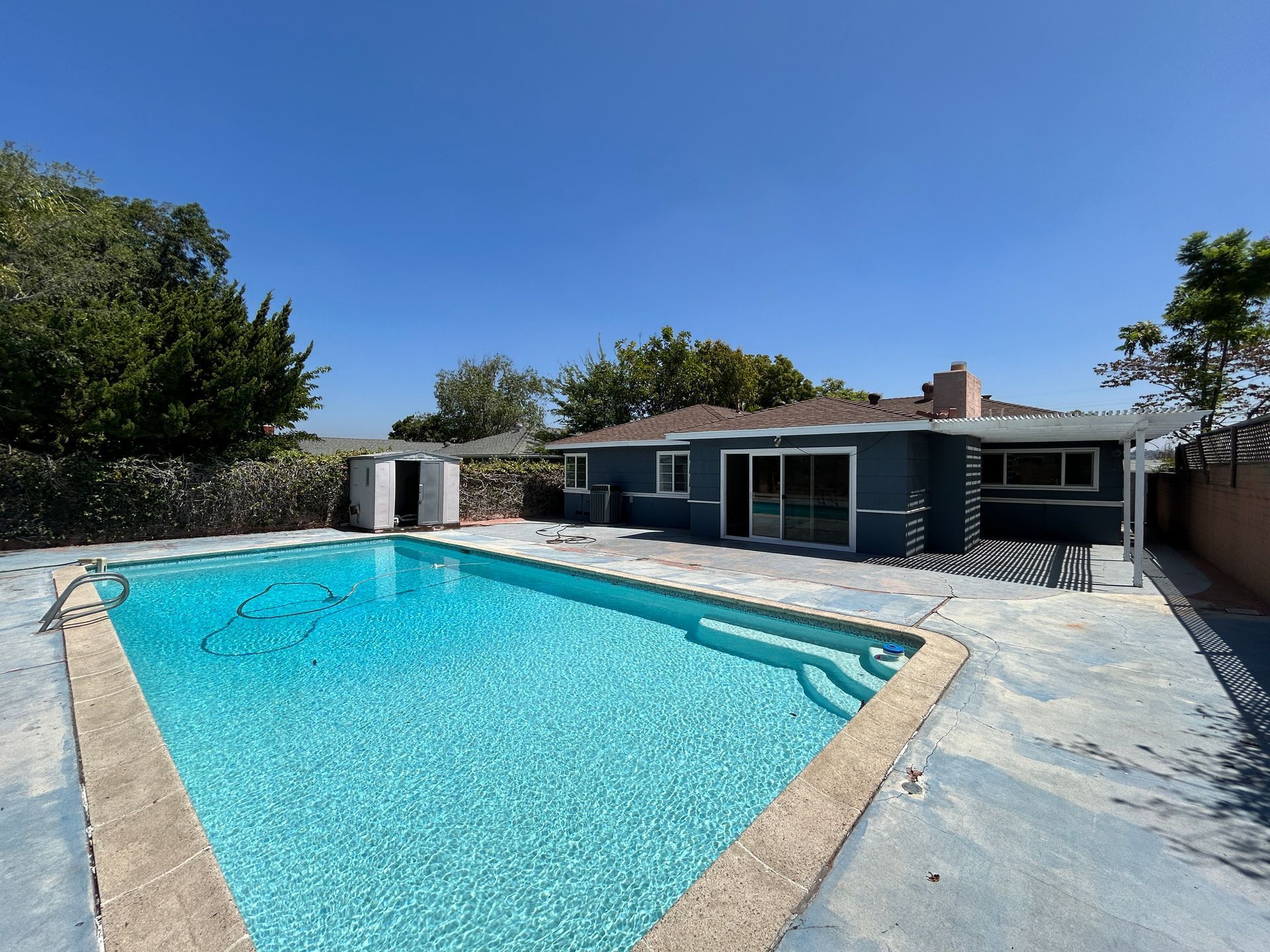 Backyard with a blue pool, a blue-gray house, and a storage shed under a bright blue sky.
