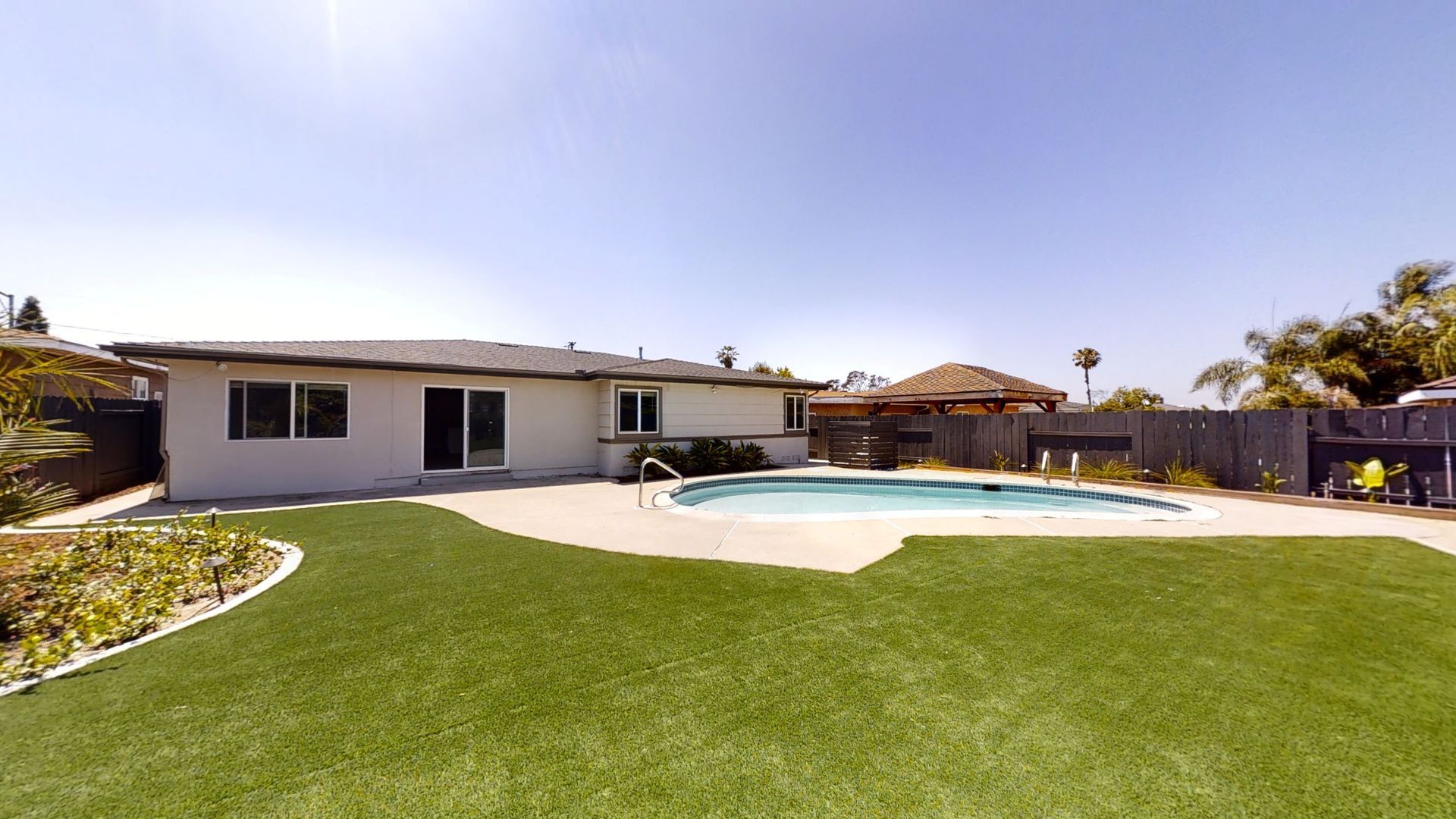 Backyard with a pool, gazebo, green grass, and a house under a clear, blue sky.