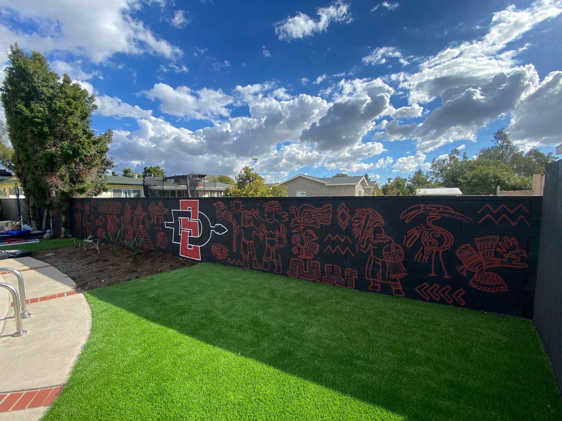 A black fence with red Aztec-themed art and San Diego State logo; green grass and blue sky with clouds.