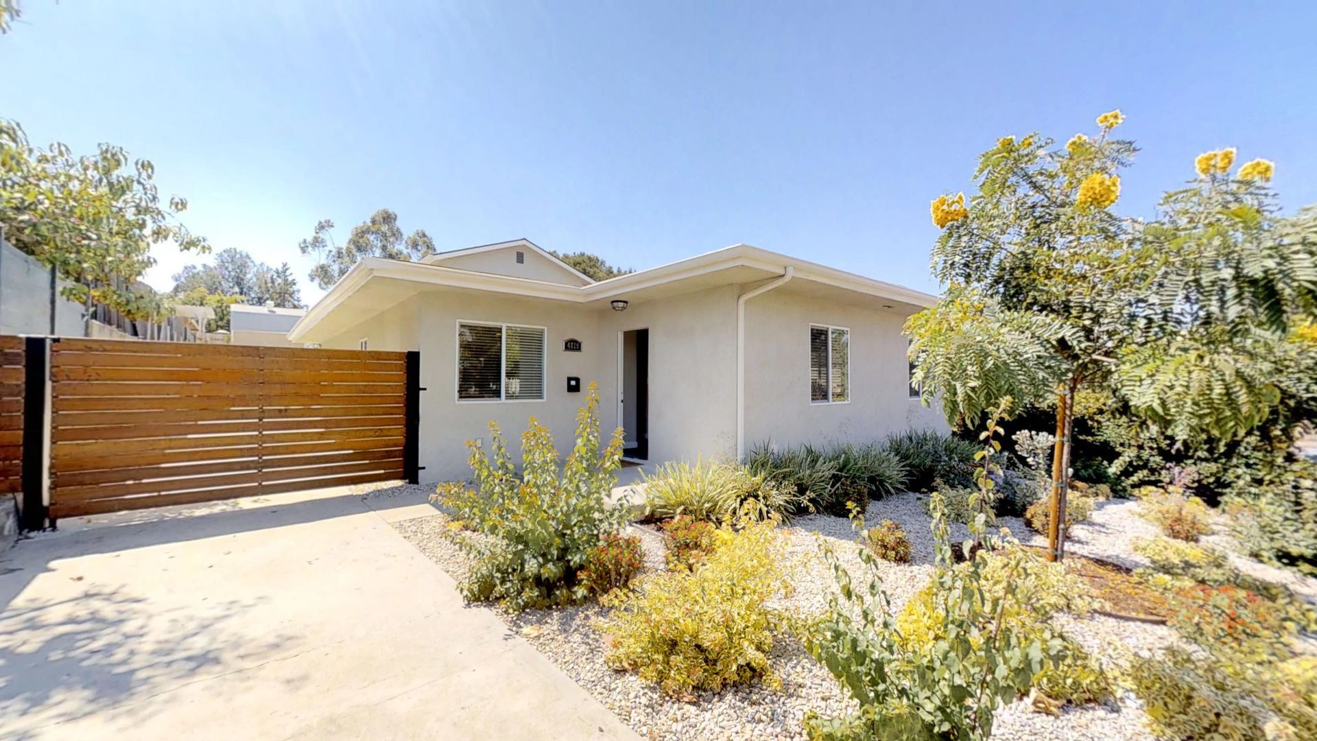 A light-colored bungalow with a wooden gate and a drought-tolerant garden. Bright sunny day.