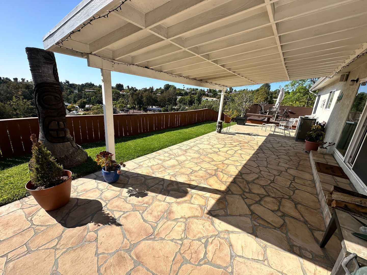 Patio with stone floor, white overhead structure, grass, and brown fence; sunny day.