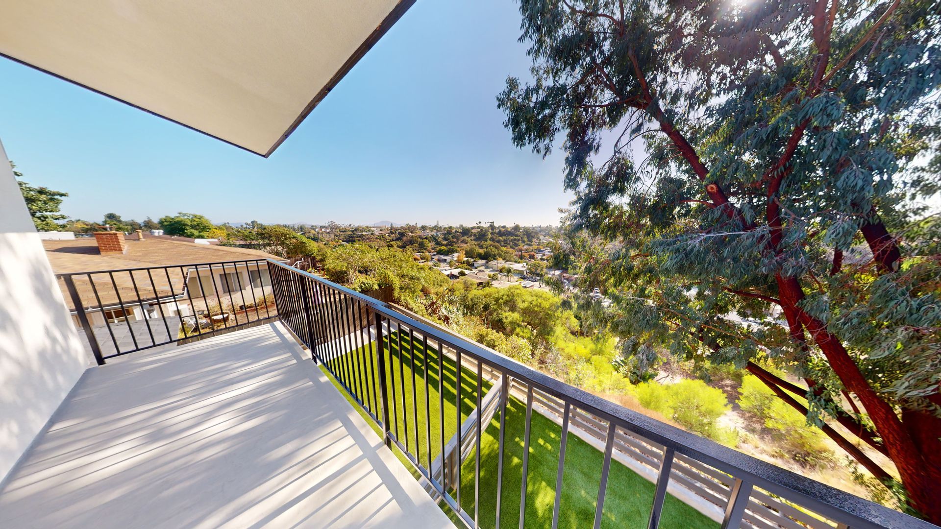 Balcony view overlooking a suburban area and trees on a sunny day. Black railing, white walls.