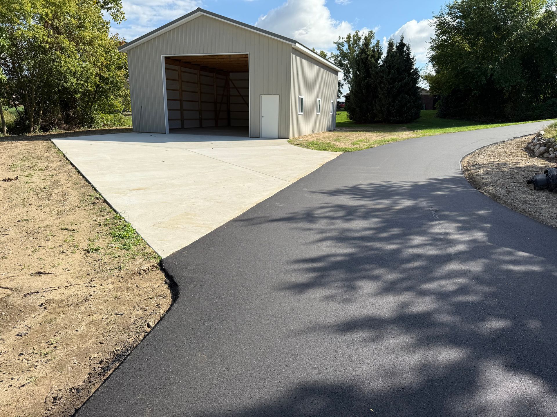 Asphalt driveway merges with a concrete pad in front of a metal building; sunny, outdoor setting.