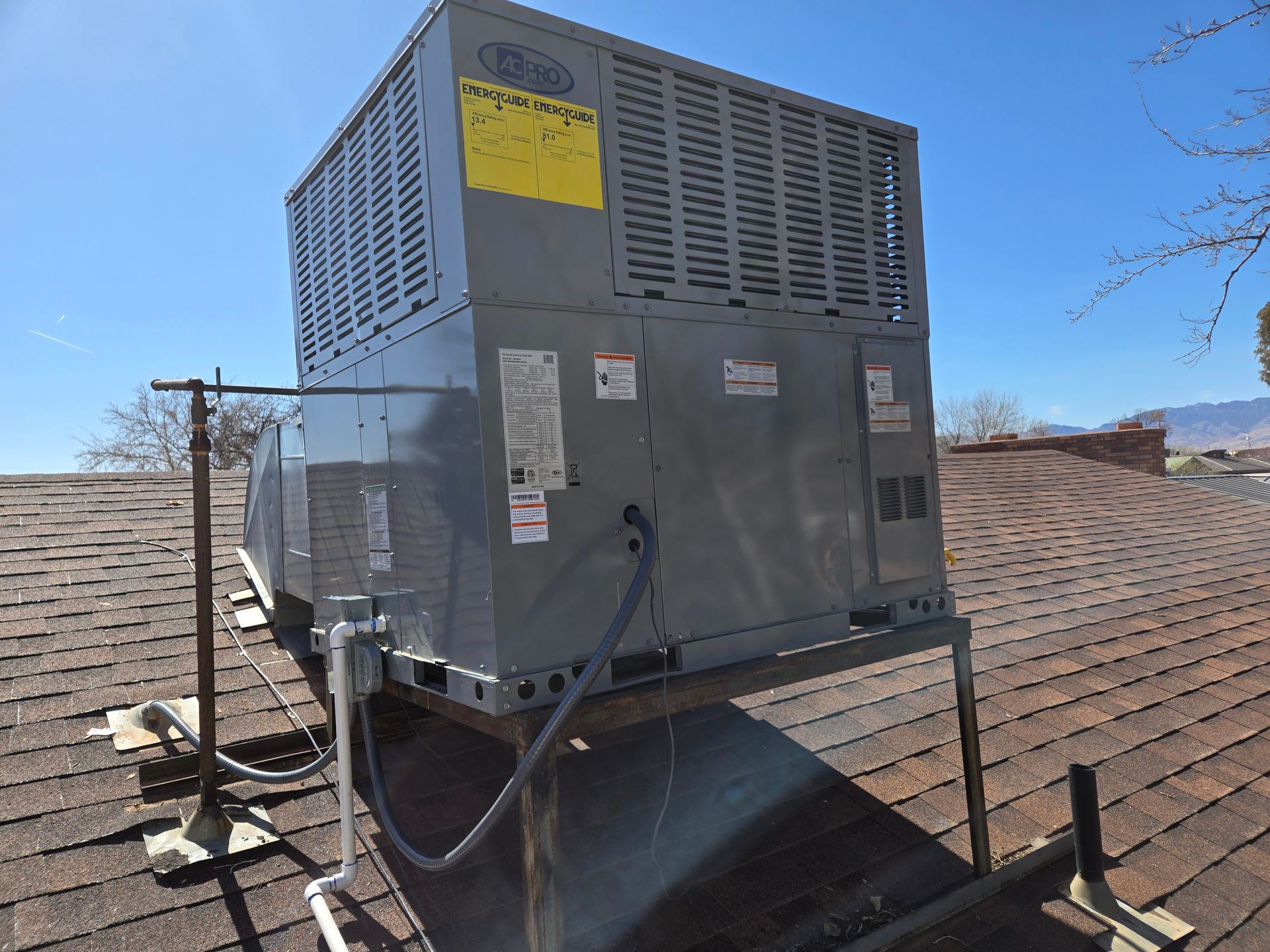 Large gray rooftop HVAC unit on a brown shingle roof against a blue sky.