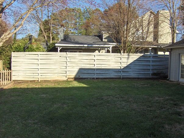 A backyard with a white wooden fence and a house in the background.