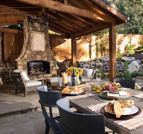 A stone masonry patio with a table and chairs and an outdoor fireplace and cedar pavilion.