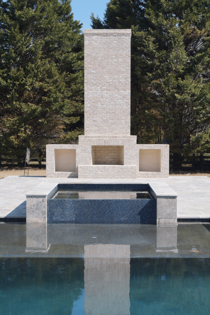 A brick fireplace sits next to a swimming pool surrounded by trees