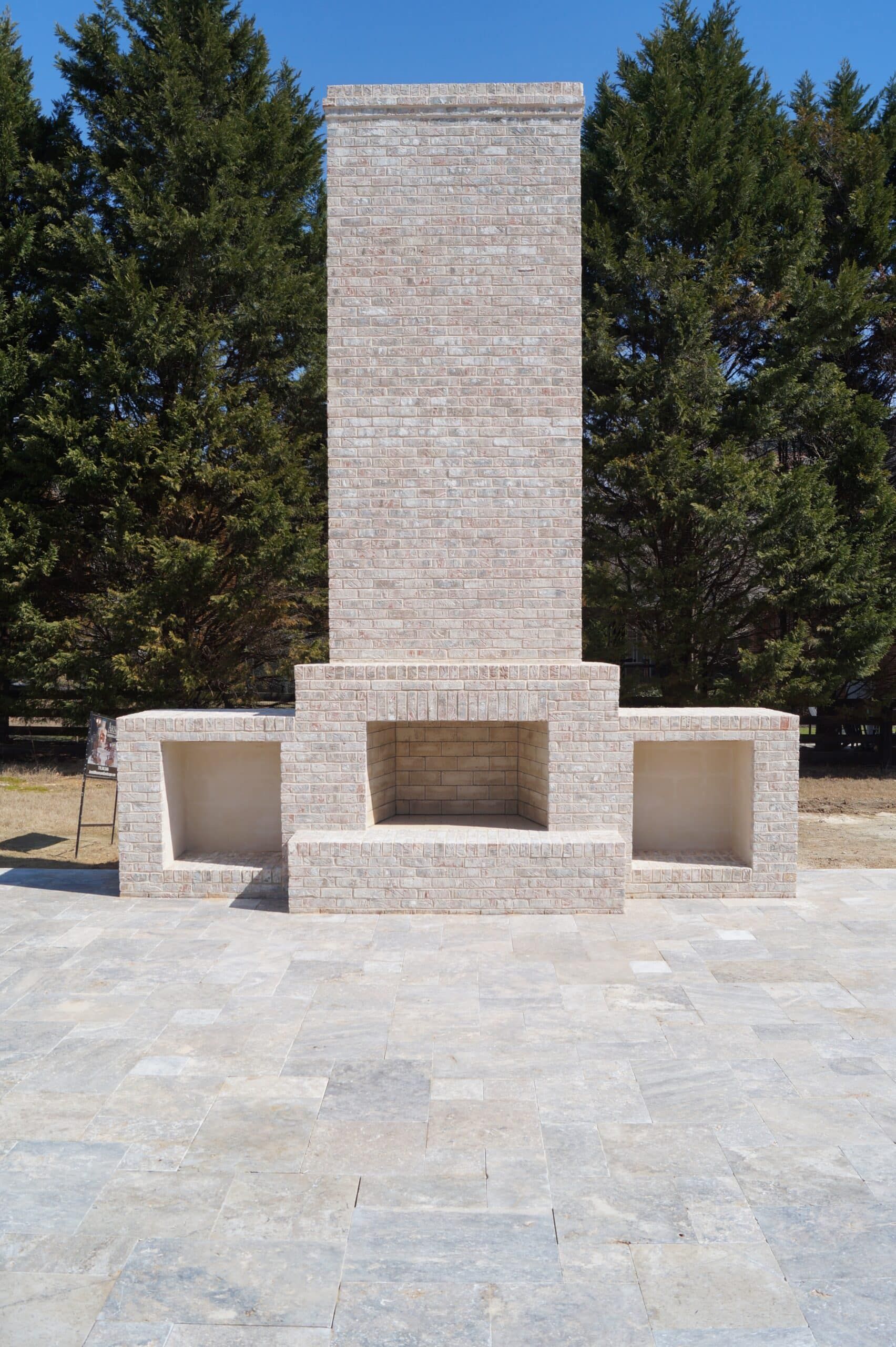 A large brick fireplace is sitting on a patio with trees in the background.