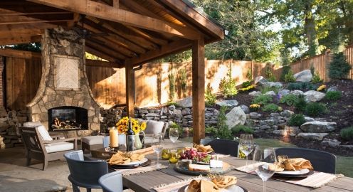 A stone patio with a table and chairs and an outdoor stone fireplace and cedar pavilion.