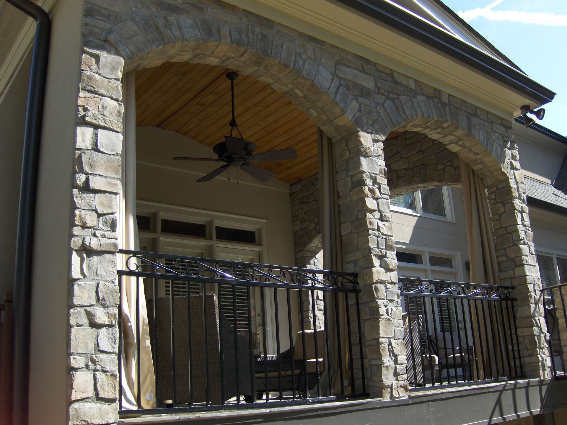 A porch with a ceiling fan on it and stone columns and arches.