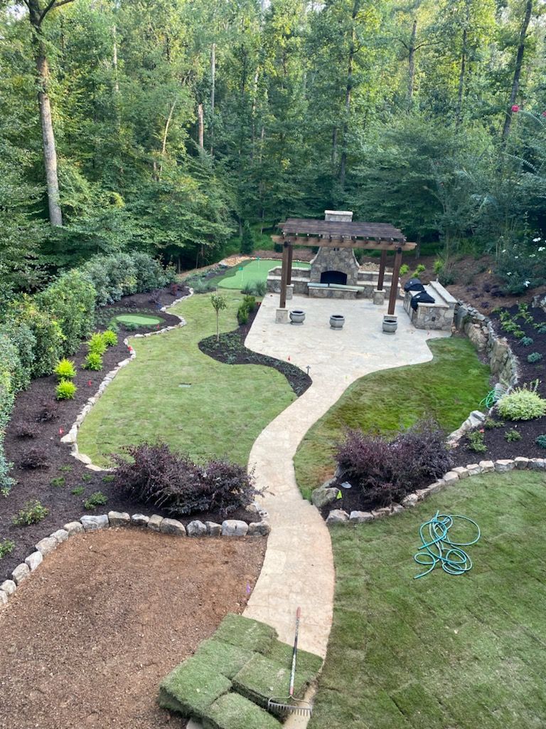 An aerial view of a lush backyard with a stone walkway, patio, outdoor fireplace, kitchen, pergola.