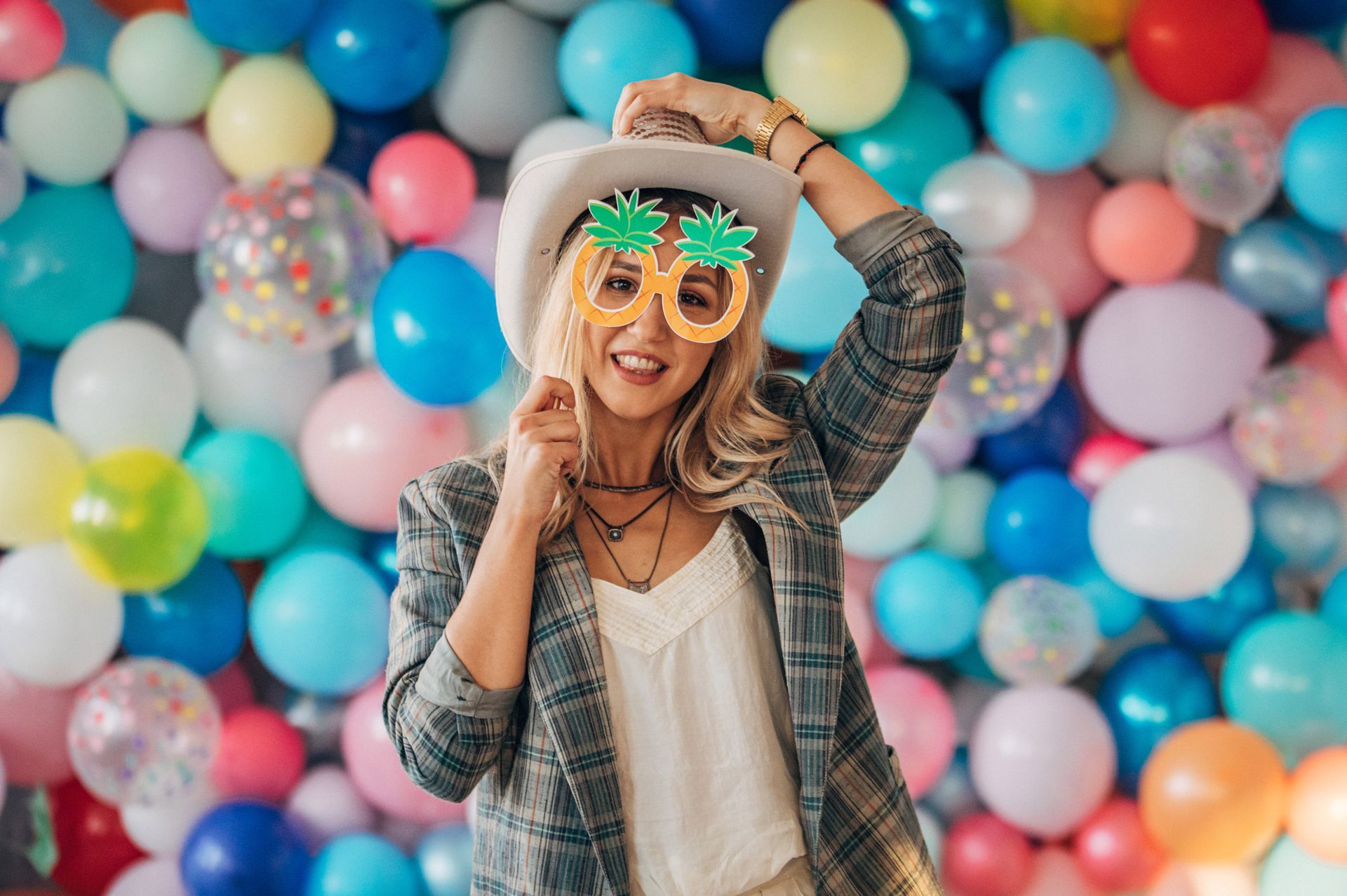 A woman wearing a hat and sunglasses is standing in front of a wall of balloons.