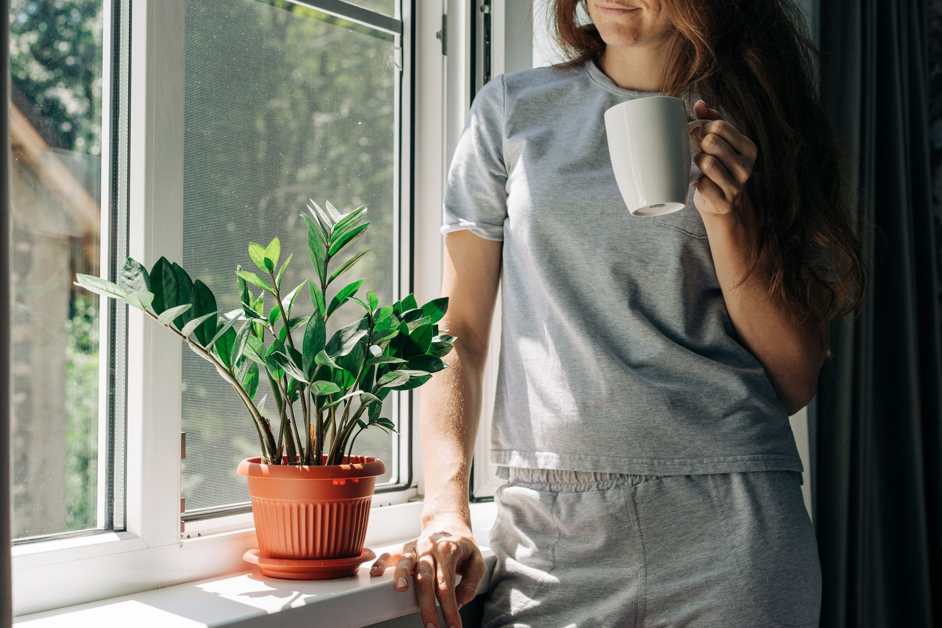 A woman holds a mug and stands by a window with a fly screen and a potted plant on the windowsill.