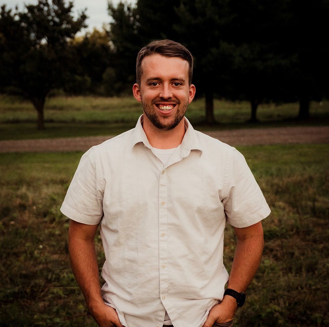 A man in a white shirt is standing in a field with his hands in his pockets.