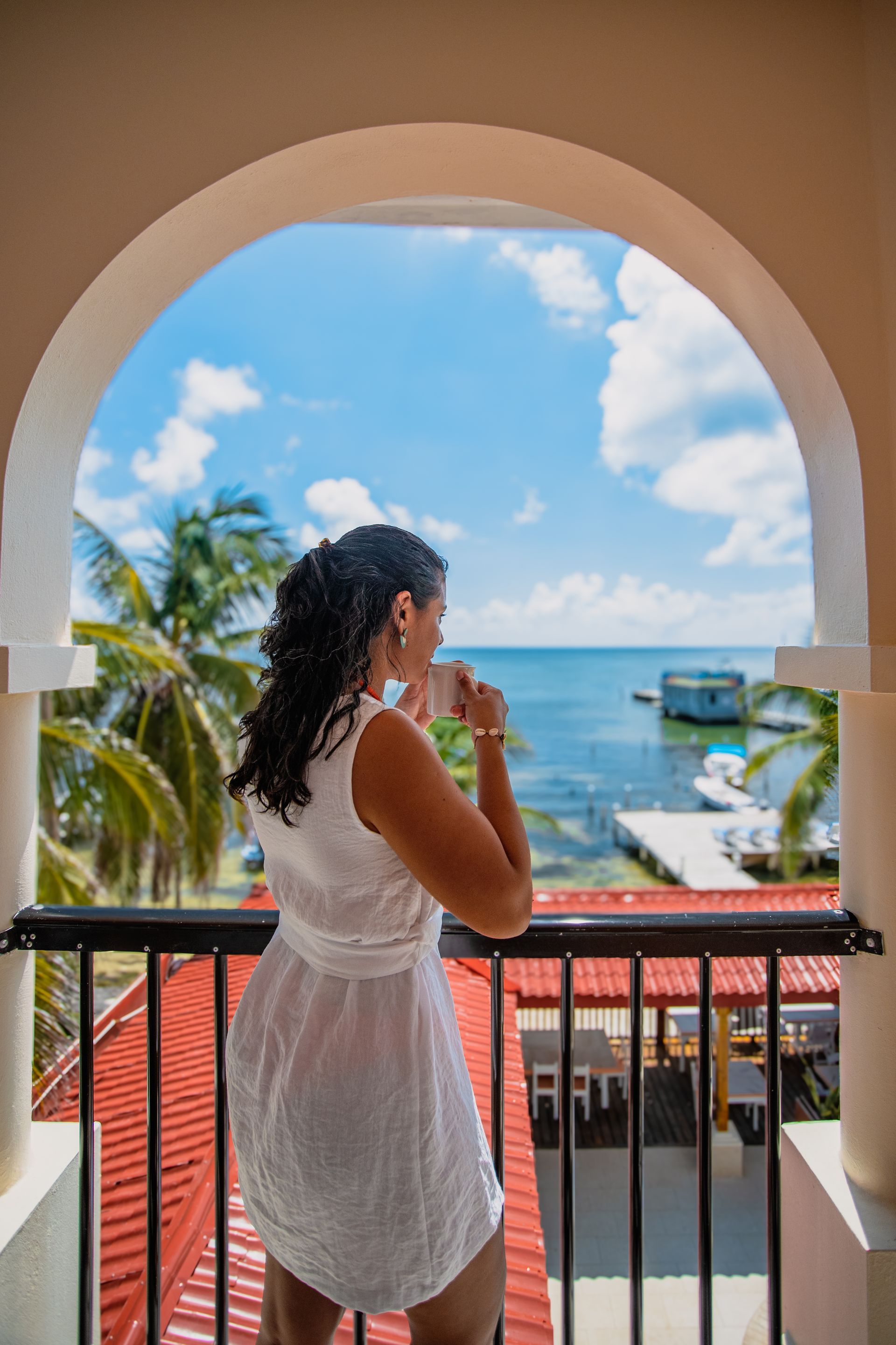 A woman in a white dress is standing on a balcony overlooking the ocean.
