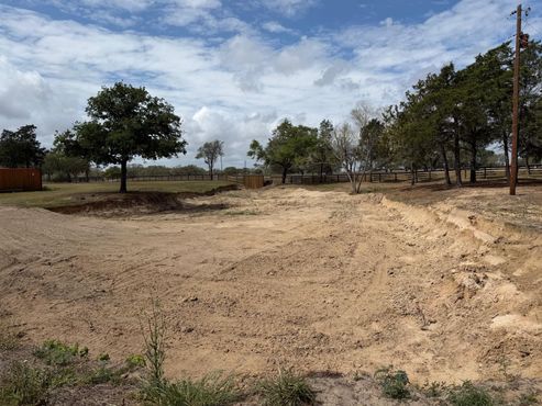A small, calm pond bordered by dry, yellowish grass under a bright, sunny sky with distant trees and a small structure.