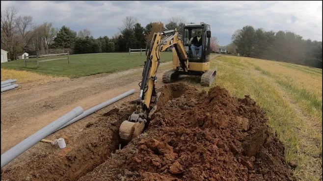 An orange plastic pipe lies inside a narrow, freshly dug trench in the dirt, with a shovel standing nearby.