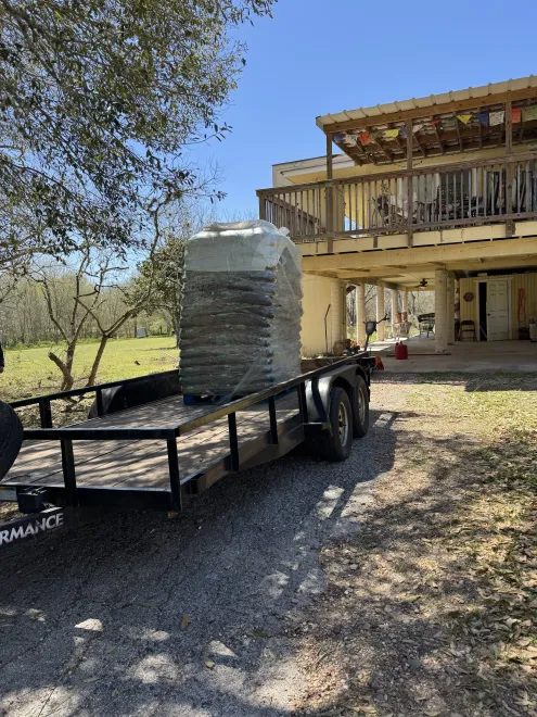 A pallet of stacked grey stone tiles sits on a flatbed trailer parked on a gravel driveway in front of a house.