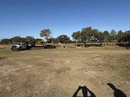 A white pickup truck pulling a trailer with a yellow excavator sits in an open, dry field under a clear blue sky.