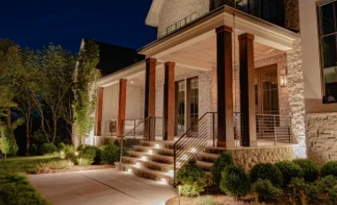 A person in a bright green hat spreads dark mulch under a wooden deck, with a rake lying on the grass in the foreground.