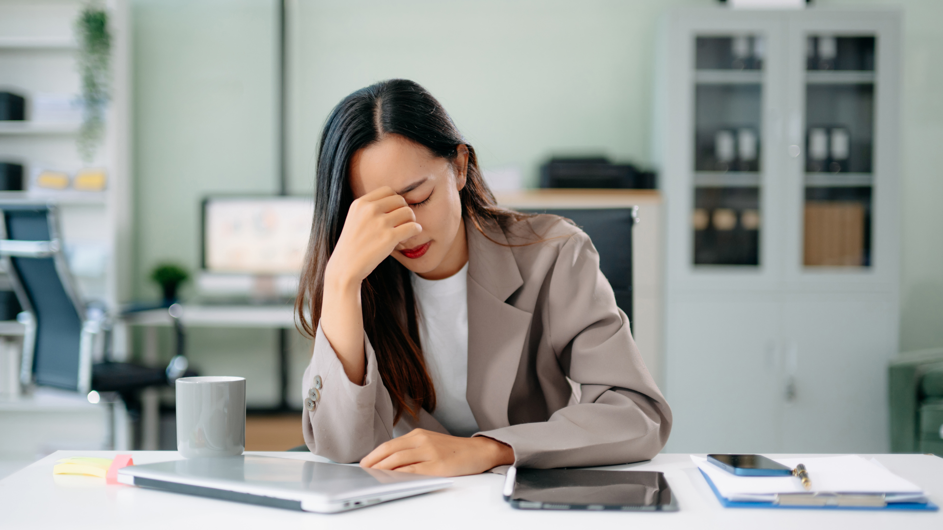 A professional sitting at a desk with eyes closed and hand on forehead, appearing stressed in an office setting.