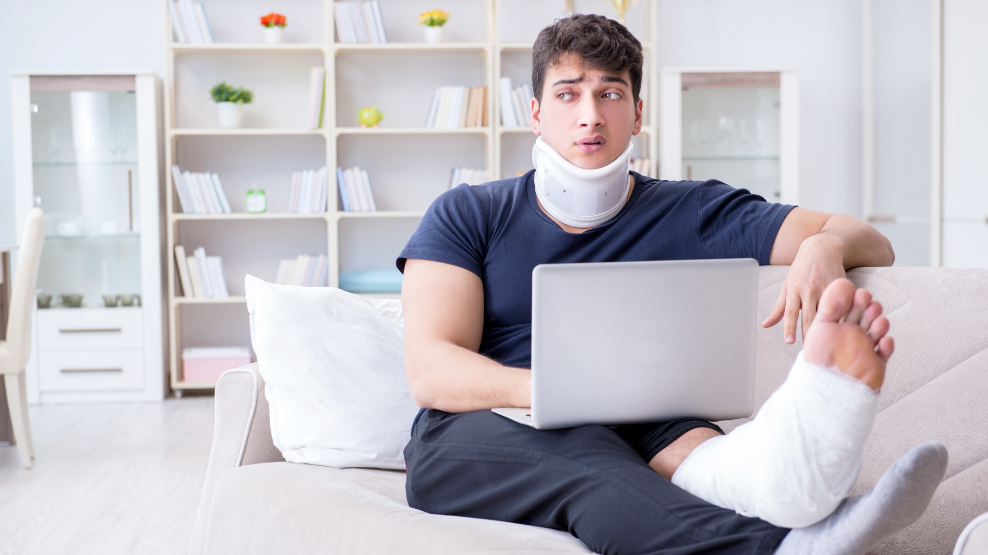 A person wearing a neck brace and with a cast on their leg sits on a sofa while using a laptop.