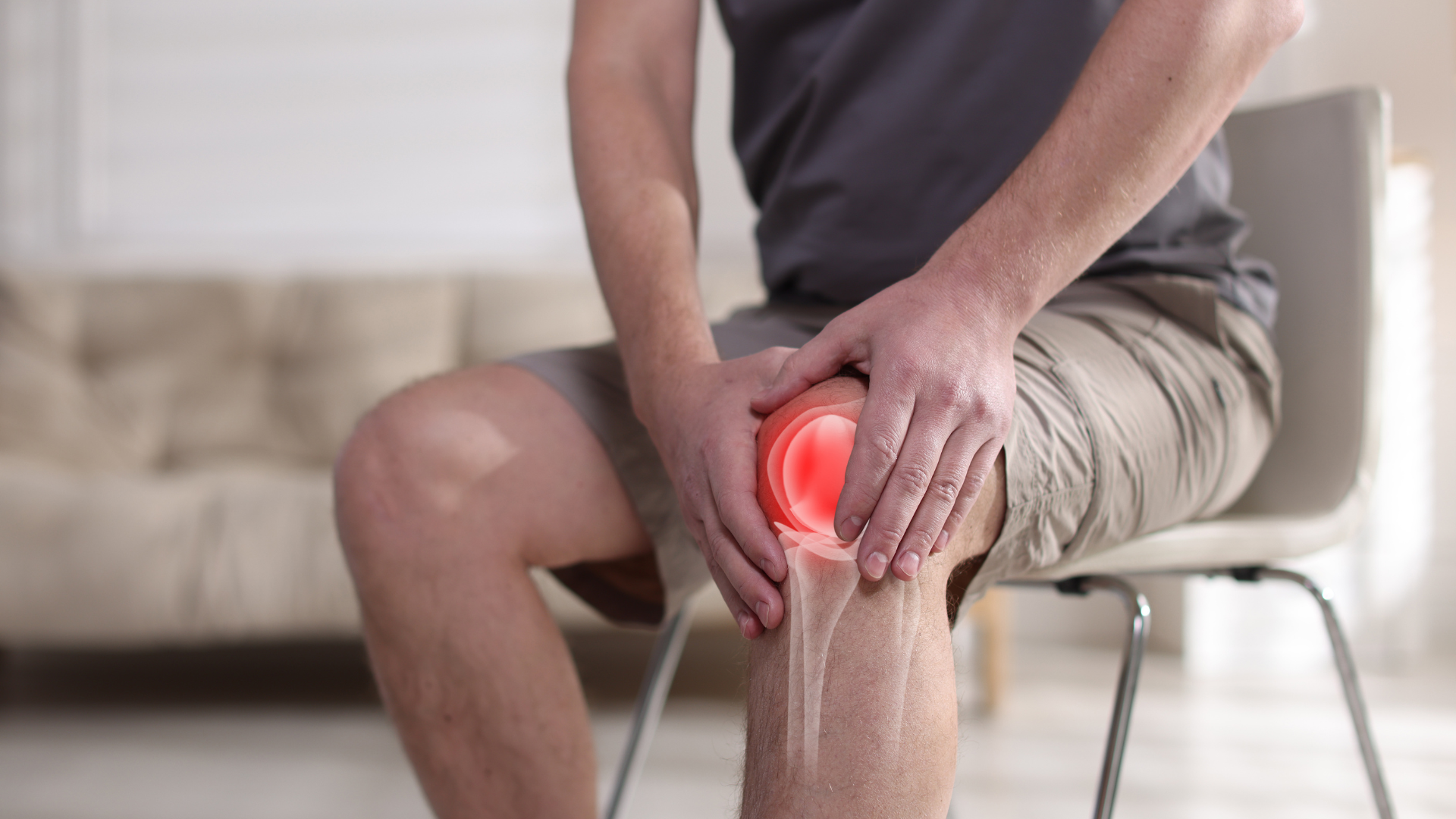 A person sits on a chair, holding their knee, which is highlighted with a red glow to indicate pain or injury.