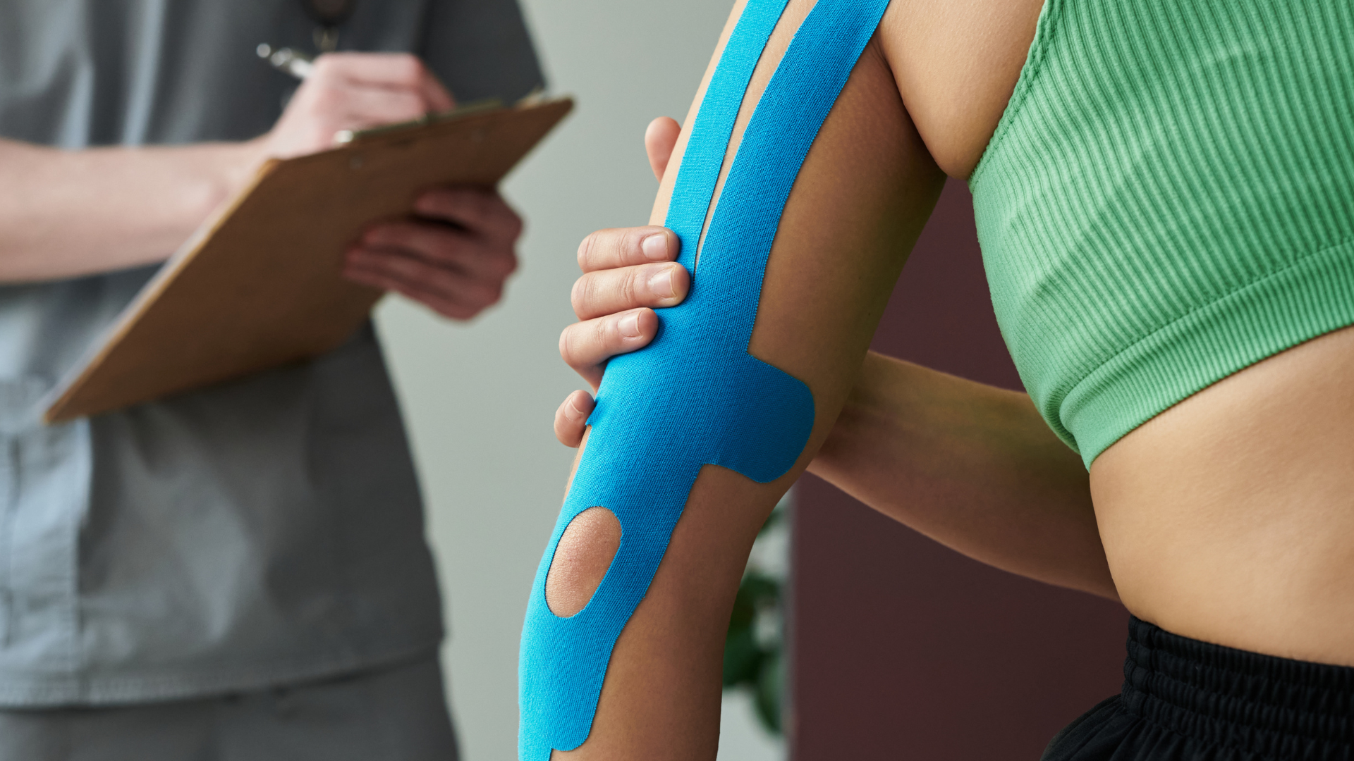 A practitioner writes on a clipboard next to a patient whose arm is supported with blue kinesiology tape.