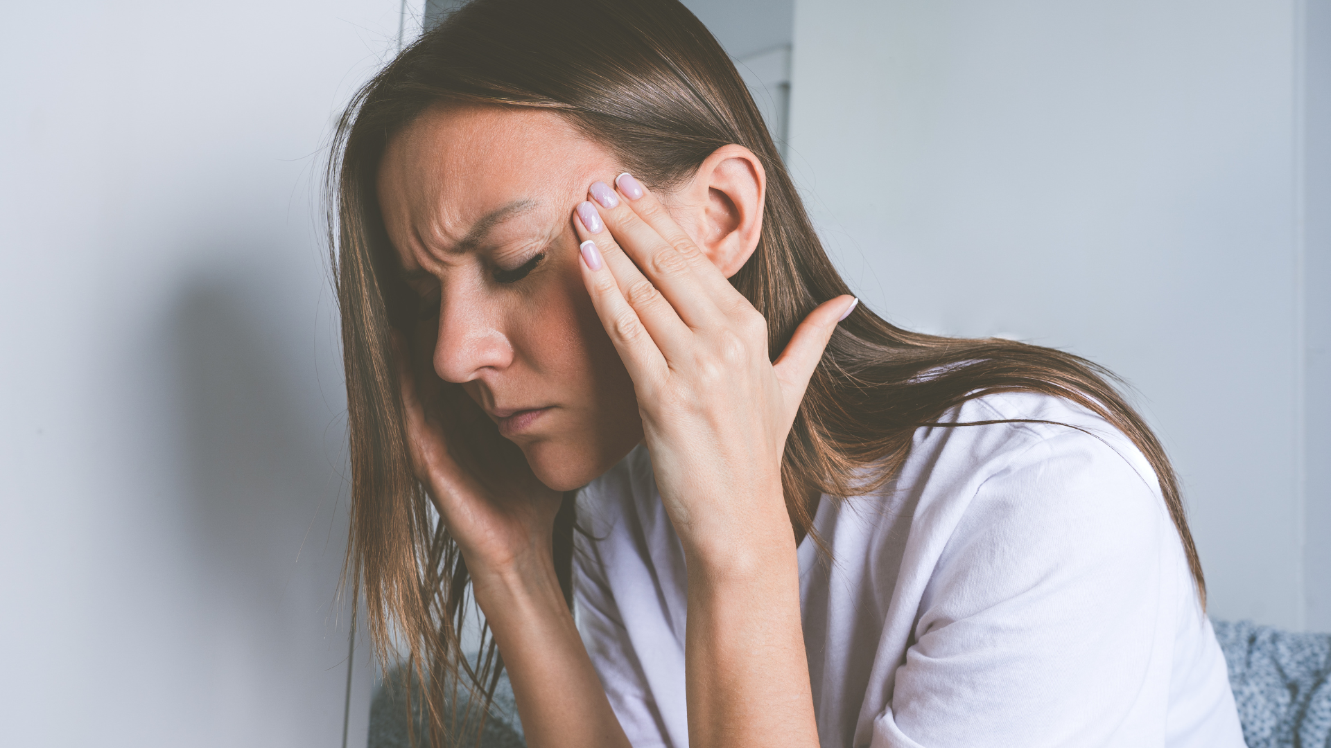 A person wearing a white shirt holds their head with both hands, eyes closed, appearing to be in pain or experiencing stress.