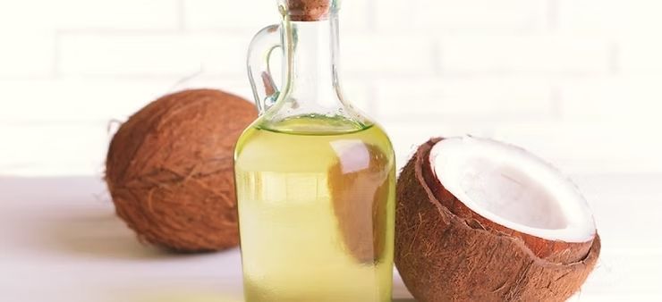 A glass bottle of light yellow coconut oil centered between a whole brown coconut and a halved coconut on a white surface.