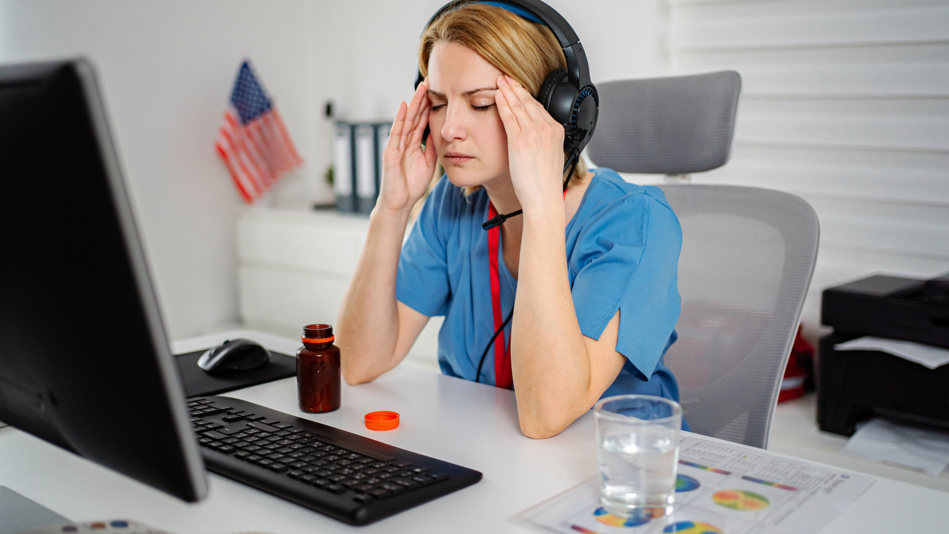 A person wearing a headset sits at a desk with a headache, hands on temples, near an open pill bottle and a glass of water.