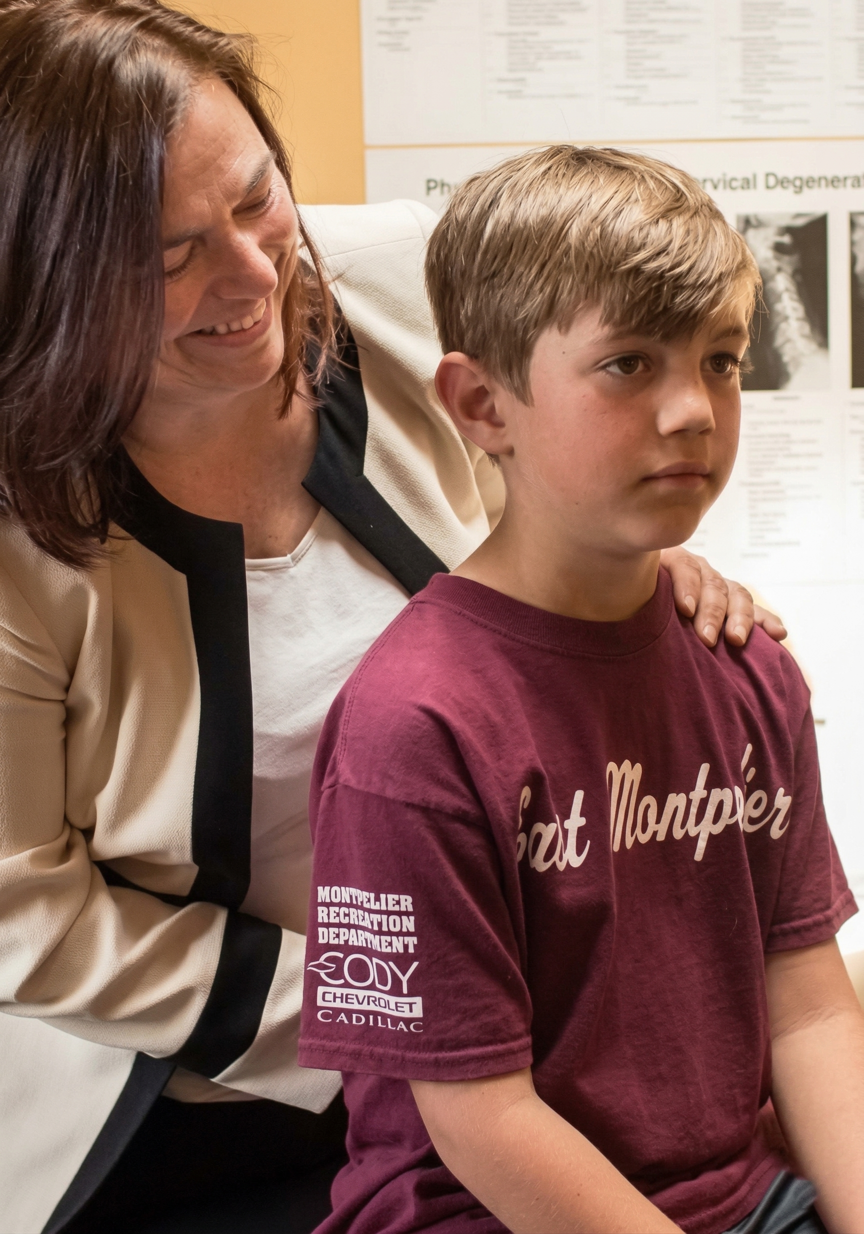 A person in a beige jacket places hands on the shoulders of a child wearing a maroon East Montpelier t-shirt.