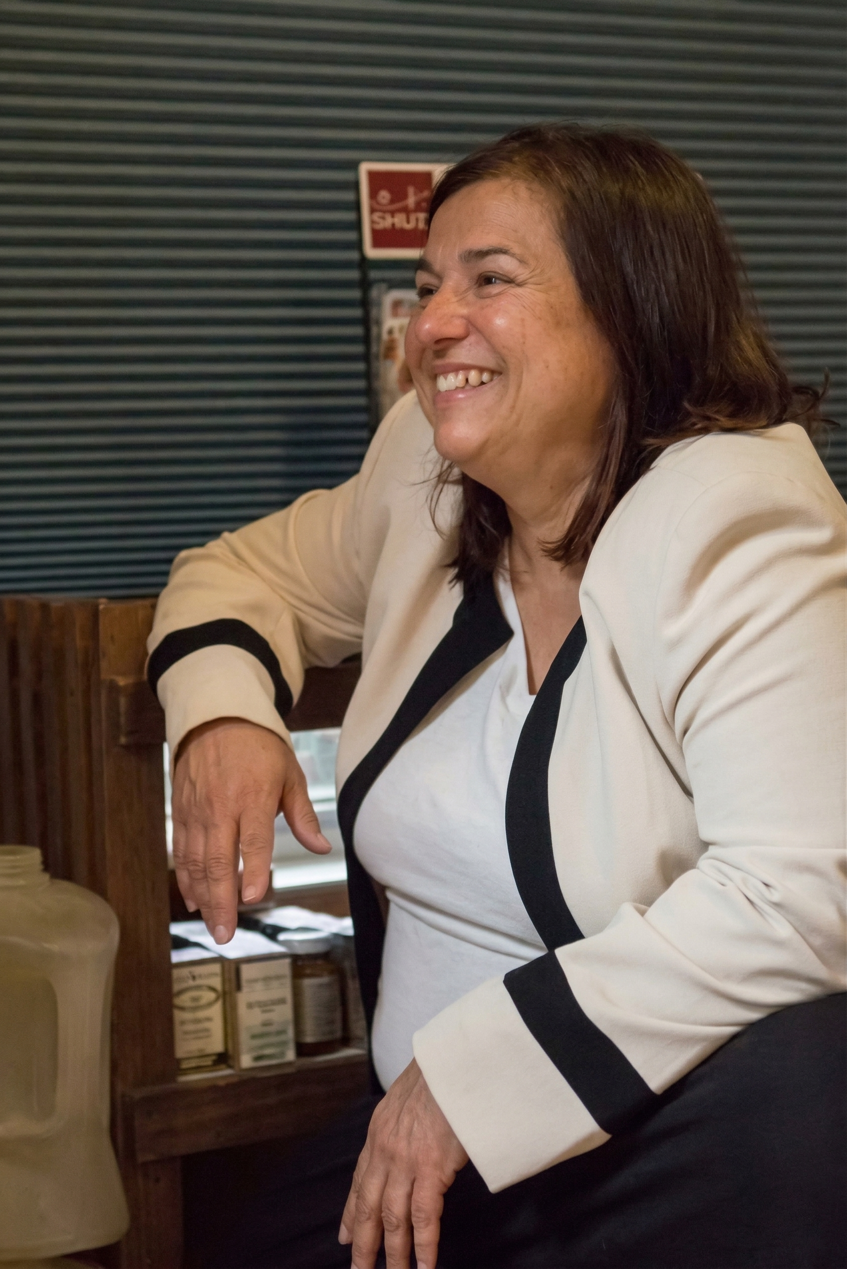 Smiling person wearing a cream-colored jacket with black trim, seated near a wooden shelf containing jars.