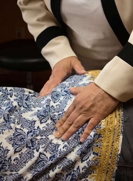 Hands press gently on the back of someone draped in a patterned blue and white fabric with gold trim.