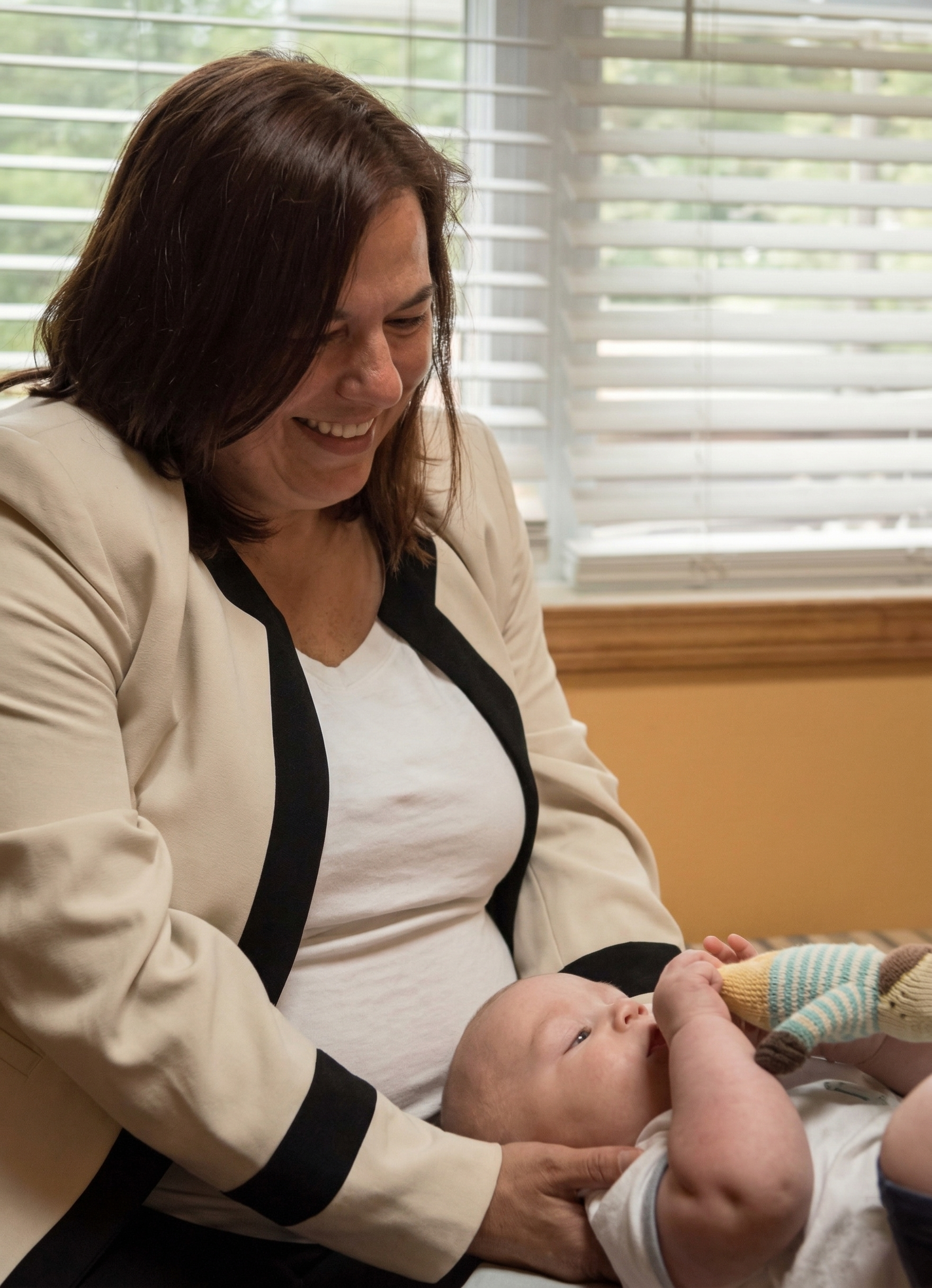 A person in a cream jacket smiles while holding a baby, who is looking at a small toy in their hands.