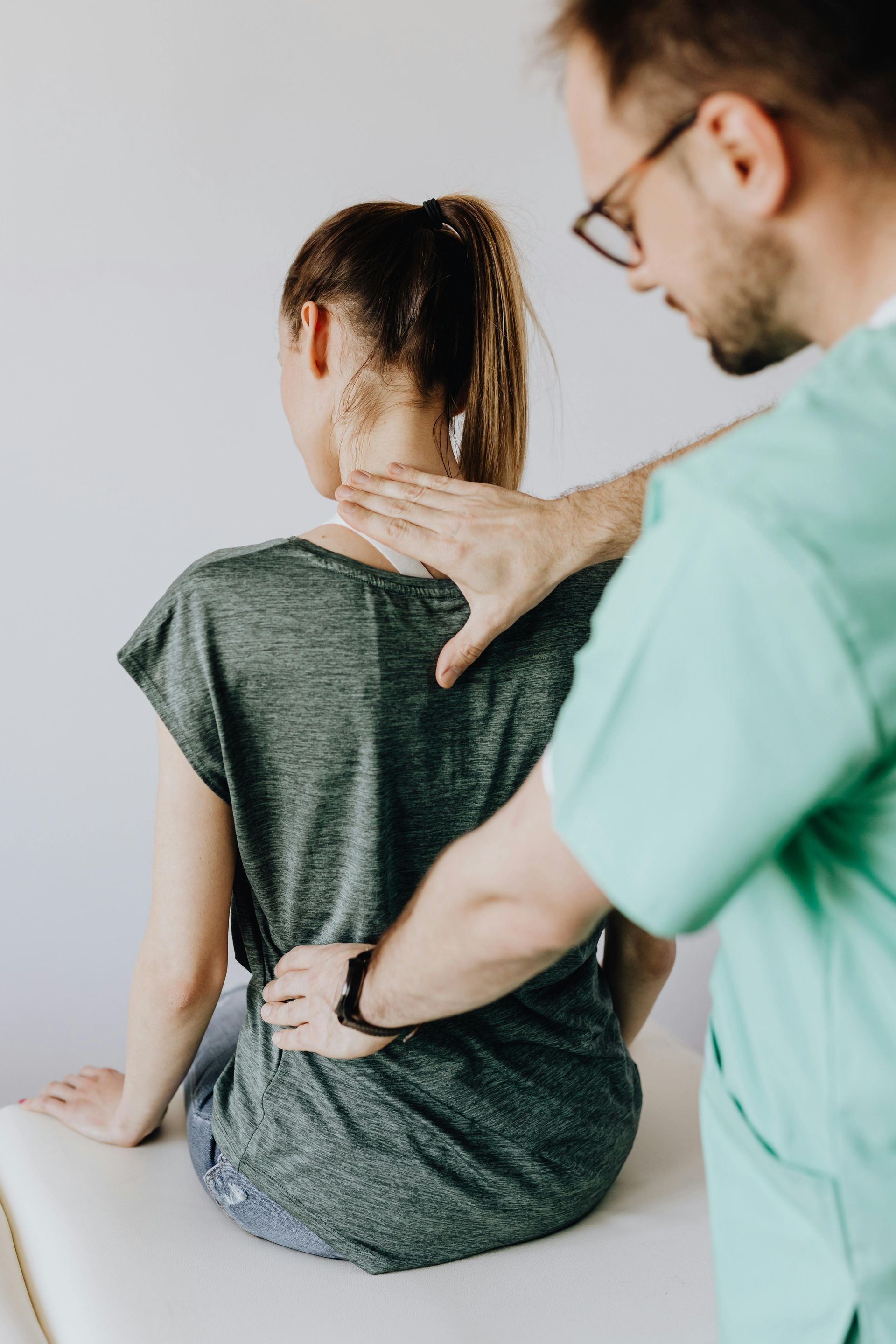A physical therapist examining a seated patient’s back and shoulder in a clinic