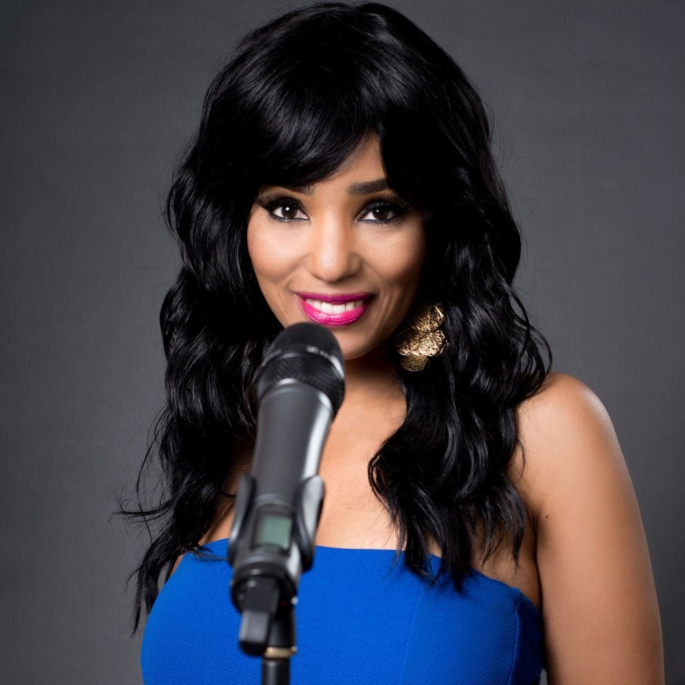 Woman with long black hair, wearing a blue dress, singing into a microphone, smiling.