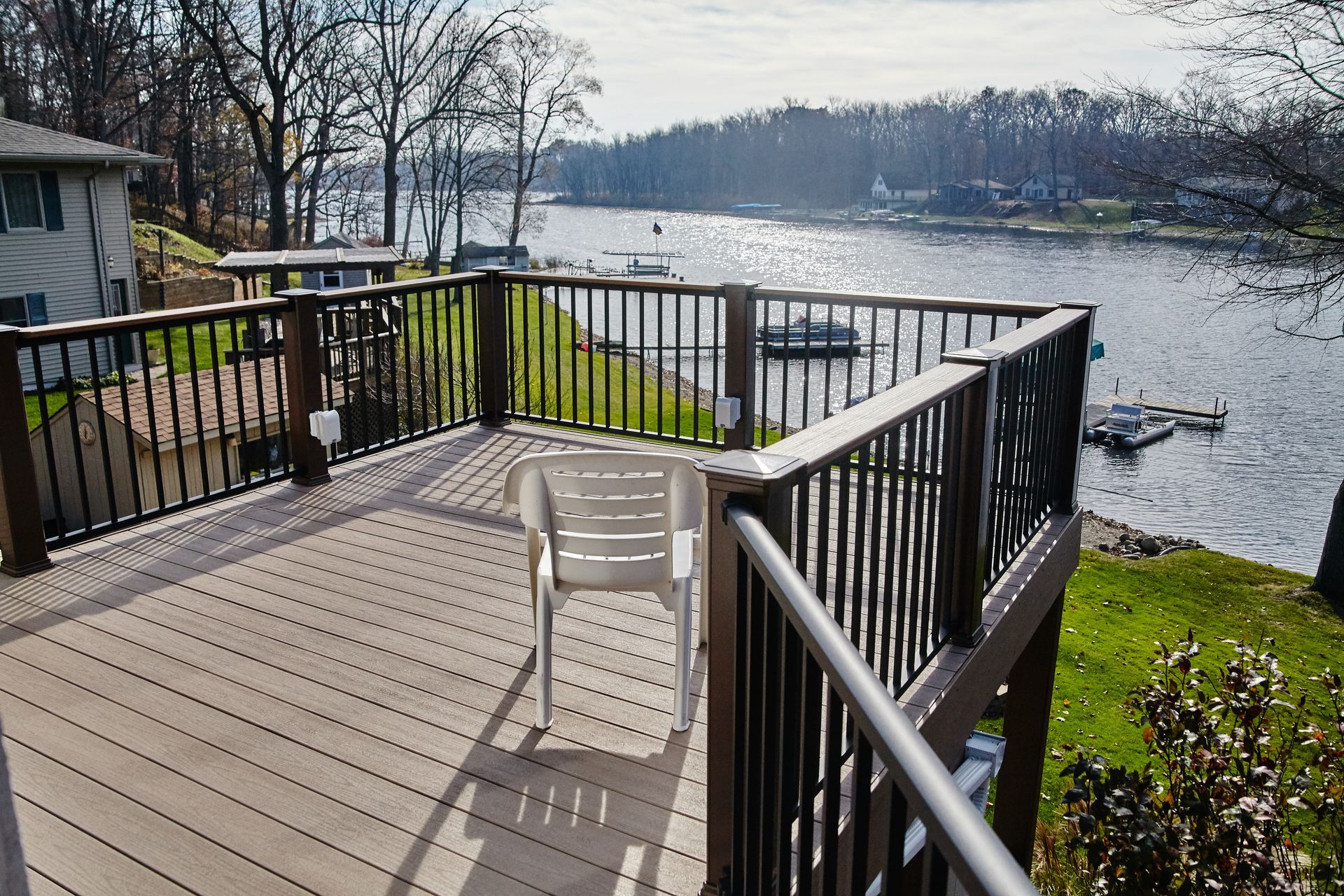 Deck overlooking a calm lake. Brown deck, black railing, white chair. Trees and houses in the background. Sunny day.